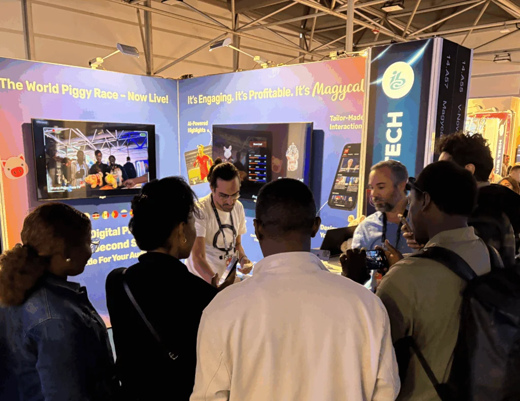 A group of people get together around Magycal's Stand at IBC 2025, to watch the World Piggy Race.